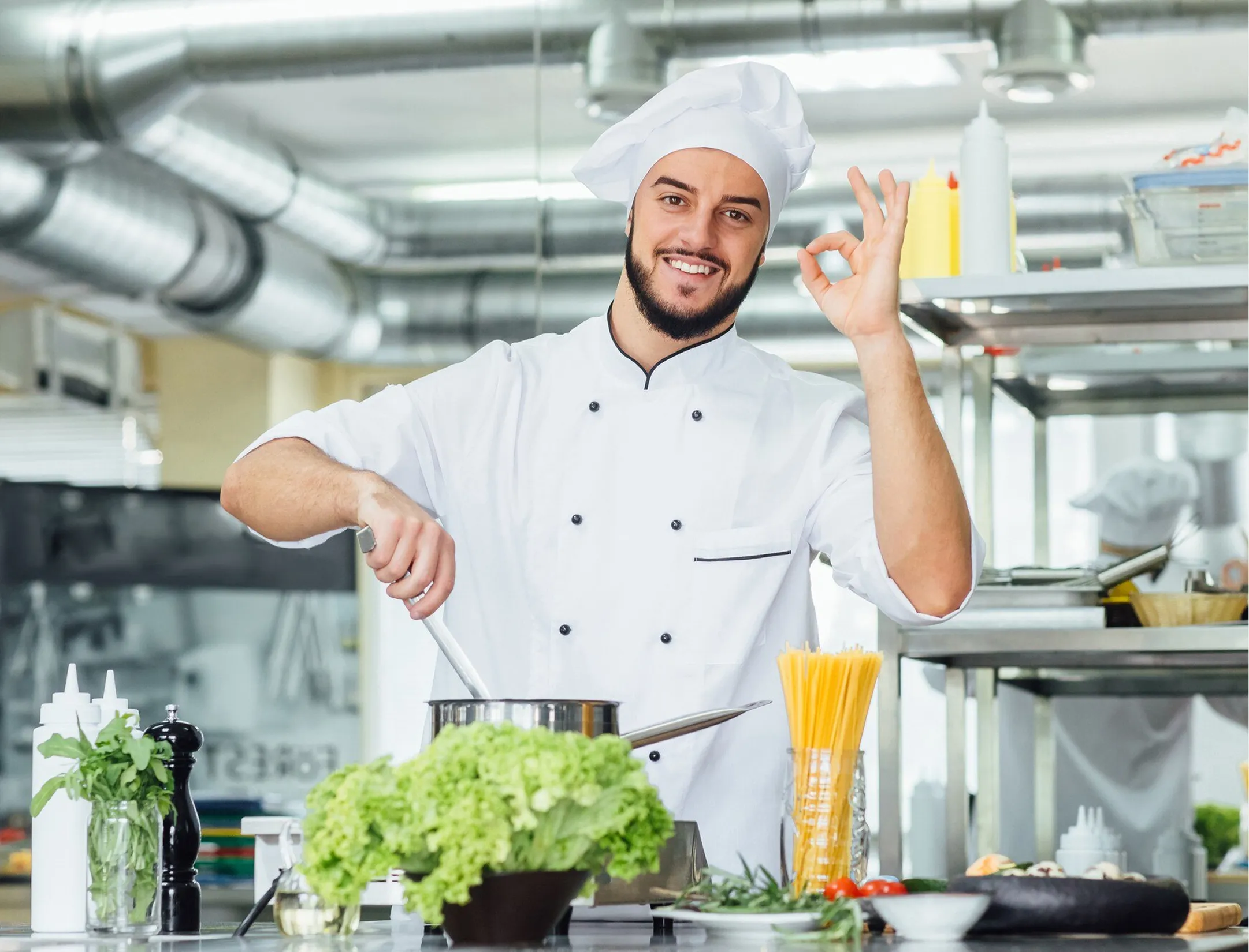 Chef in a white uniform making an 'OK' hand gesture, smiling in a kitchen setting.